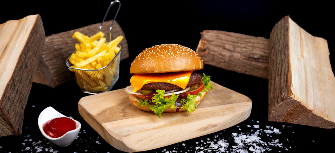 Ein Cheeseburger mit Salat, Tomate, Zwiebel und geschmolzenem Käse auf einem Holzbrett, daneben eine Portion Pommes in einem Metallkörbchen und ein Schälchen Ketchup. Im Hintergrund liegen dekorativ geschnittene Holzscheite auf schwarzem Untergrund mit grobem Meersalz verstreut.